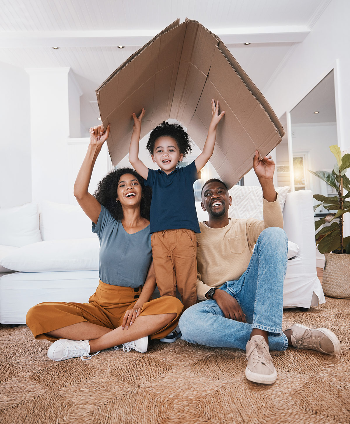 HedgeGuard Senior Benefits A joyful family in their living room, with a child wearing a cardboard box as a hat, lifted by his smiling parents, discussing senior insurance services.