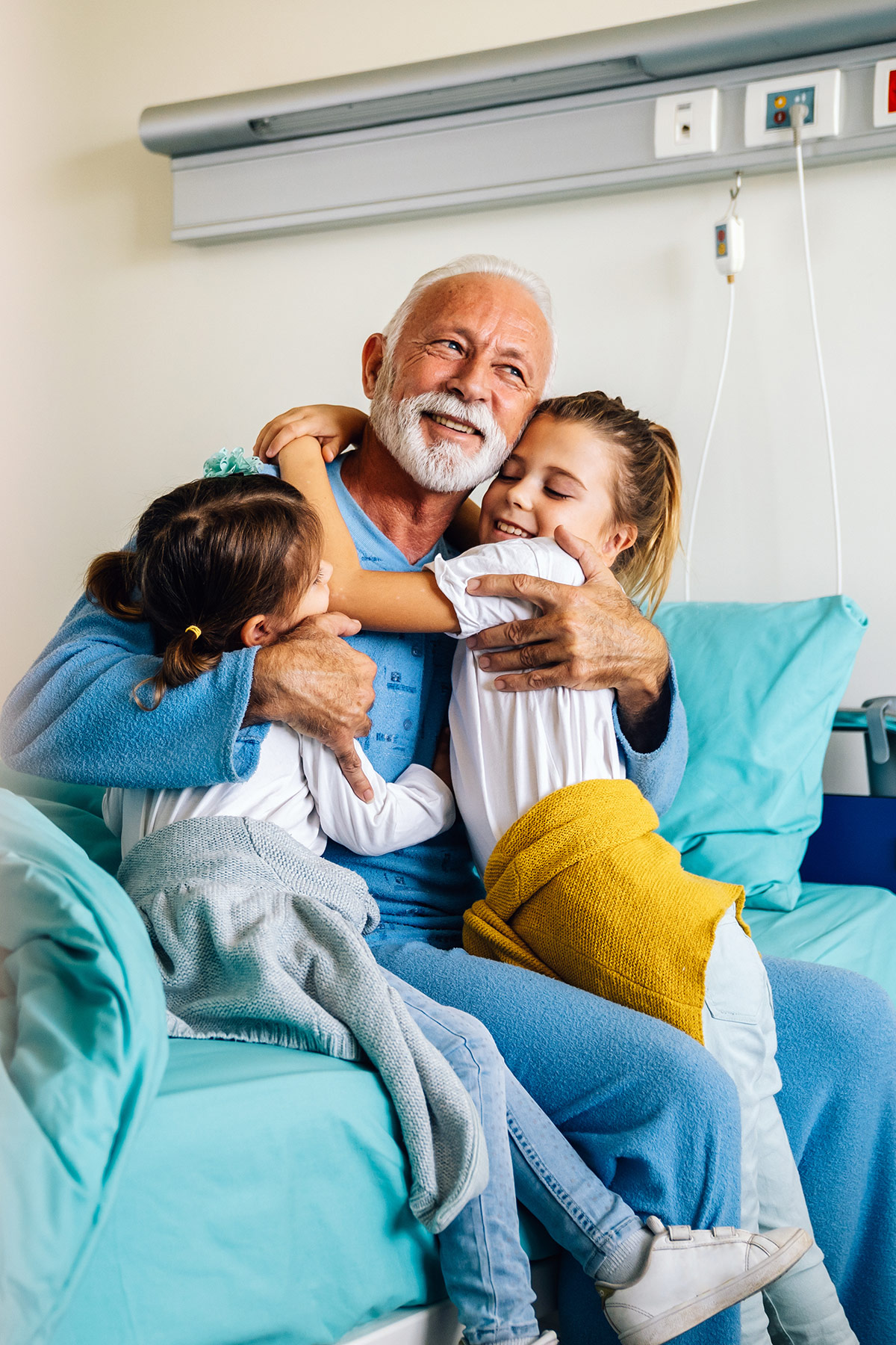 HedgeGuard Senior Benefits Elderly man with a beard smiling and hugging two young girls in a hospital room, illustrating a joyful moment supported by Senior Insurance Services.
